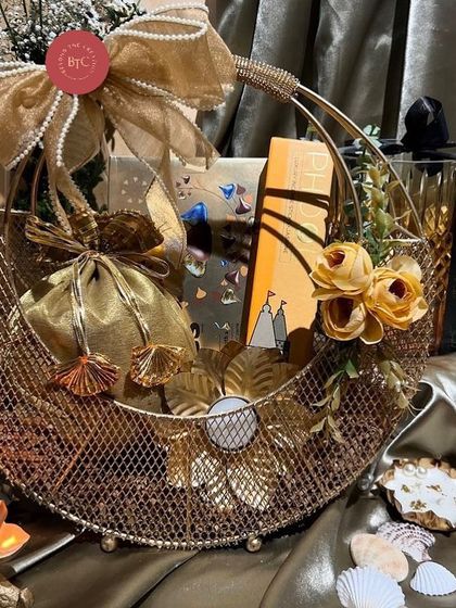 An opulent gold wire basket hamper for Diwali, filled with goodies and decorated with artificial flowers and shell-themed diyas.