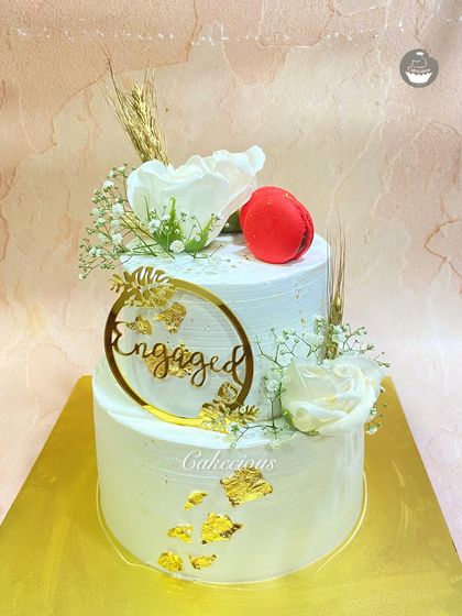 An elegant two-tier engagement cake. The simple white design is accented with gold leaf, white roses, and a single red macaron for a pop of color.