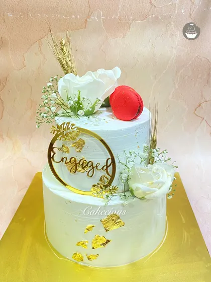 An elegant two-tier engagement cake. The simple white design is accented with gold leaf, white roses, and a single red macaron for a pop of color.