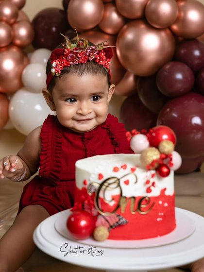 A beautiful setup for a first birthday, with a red and white cake and a stunning balloon arch in shades of rose gold and burgundy.