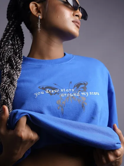 A close-up on a blue graphic tee, focusing on the "you drew stars around my scars" text. The model's cool sunglasses and braids complete the trendy, modern look.