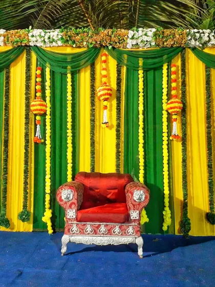 A simple stage decoration suitable for various functions like a pooja or a small ceremony. It features a green and yellow backdrop with hanging floral strings and a traditional red chair for the main guest or idol.