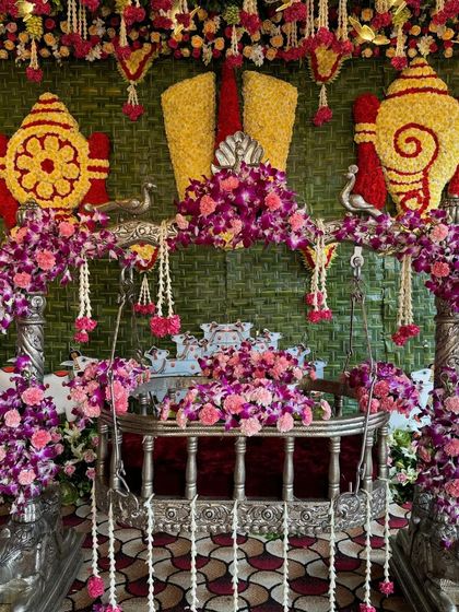 A grand naming ceremony stage featuring a silver cradle decorated with purple orchids. The backdrop has traditional shankha and chakra floral motifs.