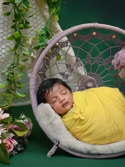 A newborn wrapped in yellow, sleeping in a purple dreamcatcher swing. The green background makes the colors stand out.