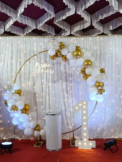 A classic and elegant first birthday setup. A golden ring is decorated with white and gold balloons, paired with a marquee number '1' and a fairy light curtain backdrop.
