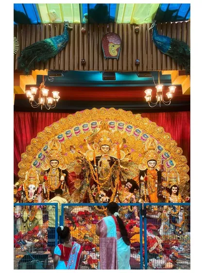 Devotees gather before a grand Durga idol, framed by peacock feather decorations, offering their prayers.