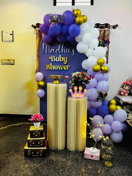 An intimate and stylish baby shower corner setup. This decoration features a purple backdrop, a white and purple balloon garland with gold accents, and elegant fluted plinths for placing cakes or gifts.