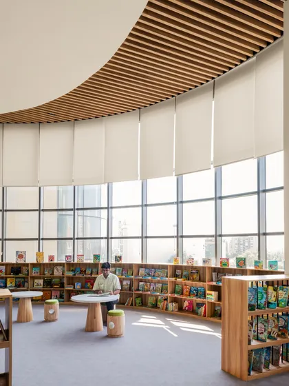 A quiet reading corner in the Vedanya School library, flooded with natural light from floor-to-ceiling windows. The curved bookshelves follow the building's organic form, creating a peaceful and inspiring atmosphere.