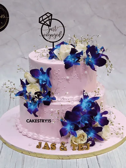 A striking two-tier mixed fruit engagement cake. The 5kg cake features a soft pink base decorated with vibrant blue orchids, white roses, and a "Just Engaged" topper.