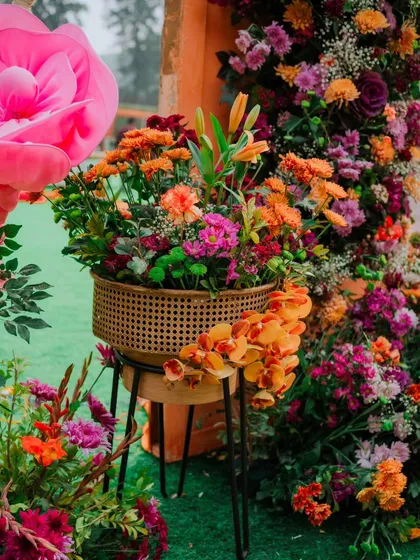 A beautiful, lush floral arrangement in a cane planter, featuring a mix of orange, pink, and purple blooms. The giant pink paper flower in the background adds a touch of whimsical scale.