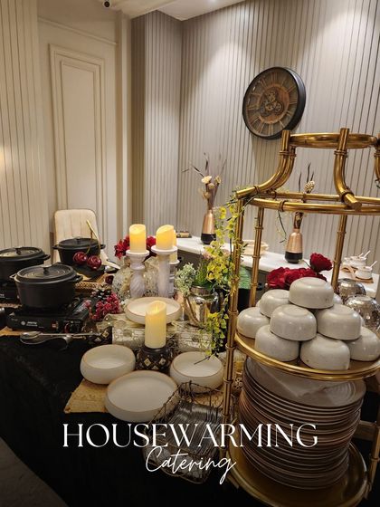 A sophisticated housewarming catering setup. The elegant buffet features gold and black accents, with a tiered stand for plates and stylish serveware.