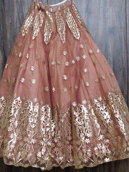 A detailed shot of the peach lehenga skirt hanging against a wooden backdrop. The image clearly shows the floral and leaf-patterned gold work on the net fabric.
