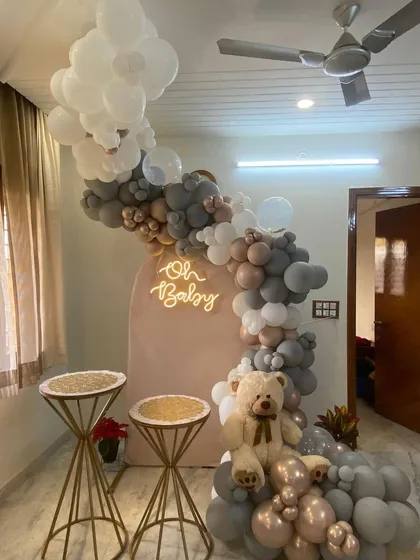 This is an example of a stylish baby shower decoration designed for a home setting. The setup includes a modern arched backdrop, an "Oh Baby" neon sign, and a sophisticated balloon arrangement in grey, taupe, and white, complete with a teddy bear.