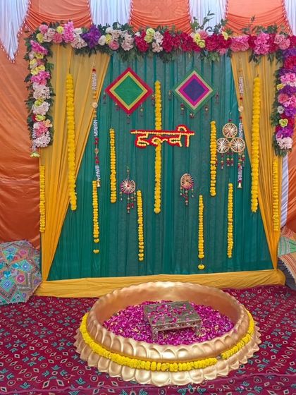 A traditional Haldi ceremony setup with a large brass urli filled with rose petals. The backdrop features green and yellow drapes, marigold garlands, and colorful hangings.