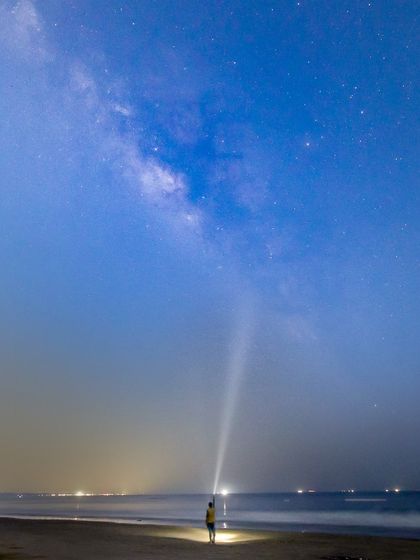A wide shot of the Milky Way from Padukere beach, captured during the blue hour just before sunrise. The single figure with a flashlight illustrates the scale of the sky.