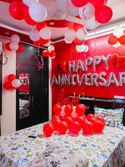 A vibrant anniversary room with bold red walls. The space is filled with red and white balloons and a large "HAPPY ANNIVERSARY" banner, creating a cheerful and celebratory environment.