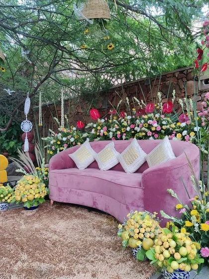 An intimate seating area for a Haldi ceremony. A curved pink velvet sofa is surrounded by arrangements of yellow flowers and lush greenery, set under a tree for a natural, garden-party feel.