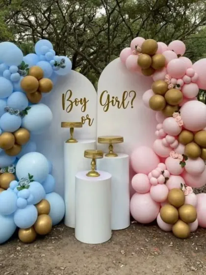 An outdoor "Boy or Girl?" gender reveal setup. The two white backdrops are adorned with blue and pink balloon garlands with gold accents, perfect for a garden party reveal.