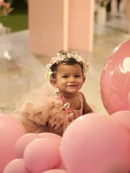 A beautiful, smiling baby girl enjoying her enchanted fairy garden-themed first birthday party, surrounded by pink balloons.