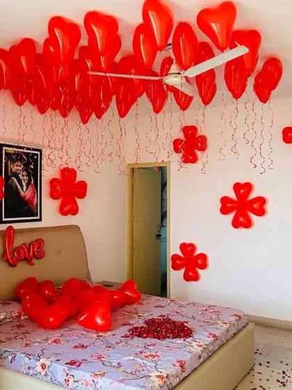 A simple yet romantic room decoration with red heart-shaped balloons floating to the ceiling. The walls are adorned with flower-shaped balloon clusters, and a 'love' foil balloon is placed on the bed.