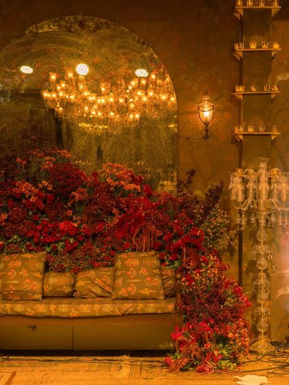 A cozy and glamorous seating area at the reception. A plush sofa is surrounded by a cascade of red flowers, with a large arched mirror reflecting the chandelier's light, making the space feel even more grand.