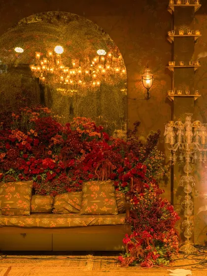 A cozy and glamorous seating area at the reception. A plush sofa is surrounded by a cascade of red flowers, with a large arched mirror reflecting the chandelier's light, making the space feel even more grand.