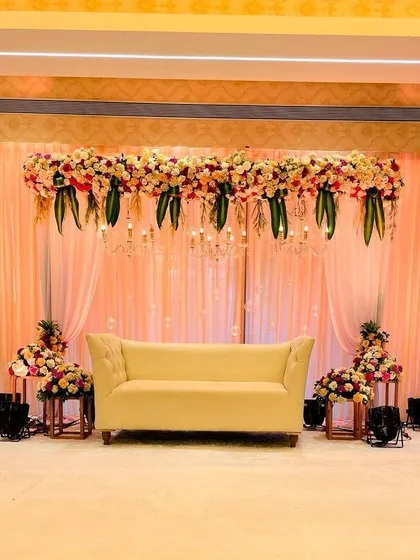 This elegant reception stage in a banquet hall uses a soft peach color palette. The design features a simple sofa, beautiful floral arrangements, and hanging chandeliers for a classic and understated look.