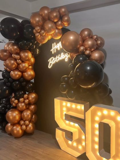 A bold and stylish 50th birthday setup featuring a black backdrop with a neon sign. A balloon arch in metallic bronze and black complements the large, light-up marquee numbers.