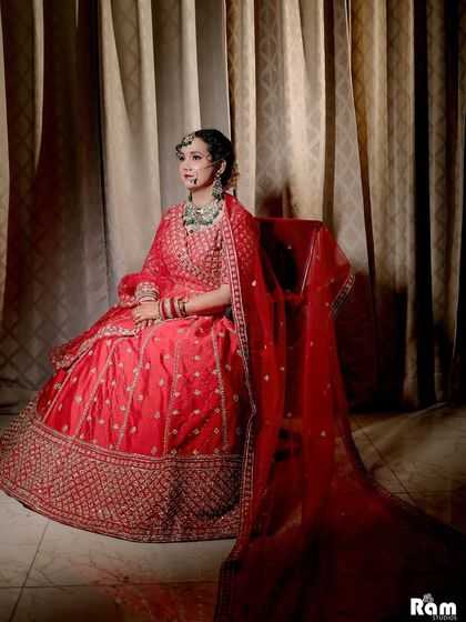 An elegant portrait of the bride seated in her red lehenga, with the soft curtain backdrop adding to the classic feel of the shot.