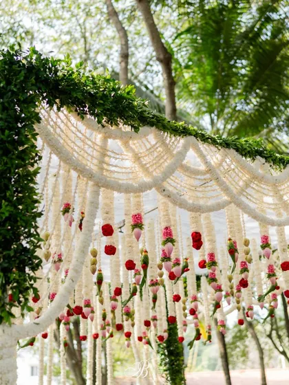 A detailed shot of the mandap's floral canopy. You can see the intricate layers of tuberose garlands and the pops of color from the hanging lotus buds, red roses, and handcrafted leaf parrots.