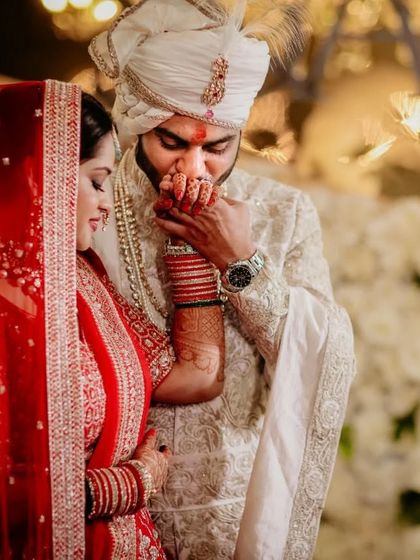 A gesture of chivalry and love. The groom kisses his bride's hand, a timeless romantic moment captured in the warm glow of the wedding lights.