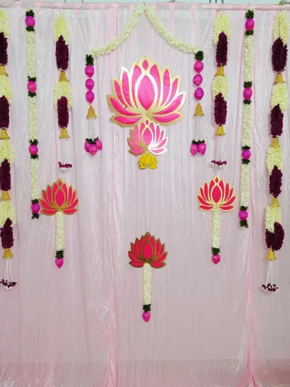 A beautiful floral backdrop for a traditional ceremony. The wall features hanging garlands of artificial flowers and golden lotus cutouts, creating an elegant and spiritual ambiance.