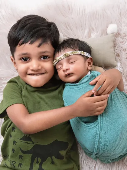 A proud big brother smiles for the camera while hugging his swaddled newborn sibling. Capturing the personalities of both children is key to a successful sibling photoshoot.