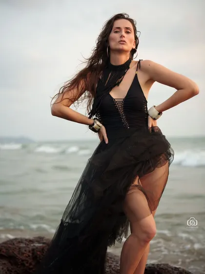 A strong, confident pose on the rocks, with the waves crashing in the background. The black outfit stands out against the natural colors of the sea and sky.