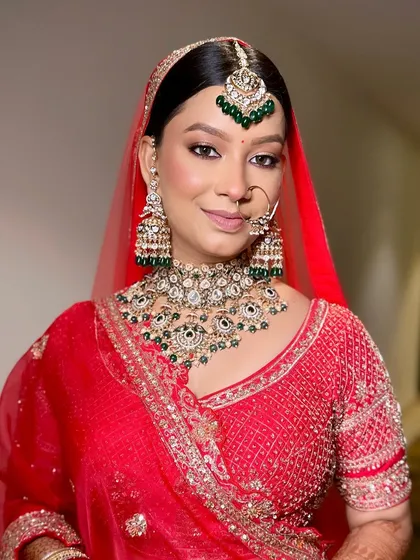 A close-up of a bride in a classic red lehenga. This shot perfectly showcases the heavy Kundan and green bead jewellery set, including the oversized jhumkas and matching maang tikka.