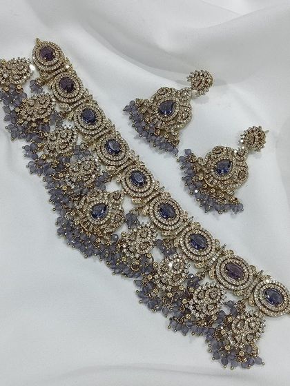 This choker set is available in multiple colors including ruby, emerald, and grey. The intricate metalwork combined with colored stones and pearl or bead droppings offers a sophisticated and royal look.