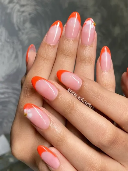 A vibrant orange French tip on almond nails. The single, small floral accent with a gold stud adds a delicate touch to this otherwise bold and summery look.