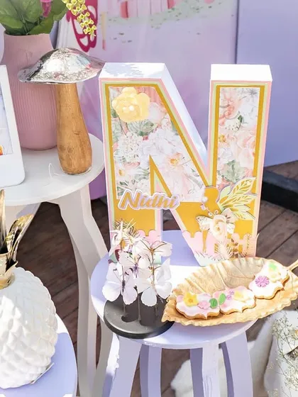 A personalized 'N' for Nidhi's fairy-themed party, surrounded by other whimsical decor elements like a pineapple vase and framed photos.