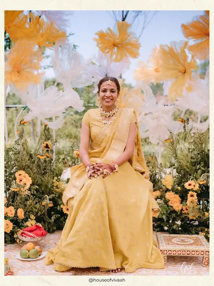 The bride, radiant in yellow, seated for her Haldi ceremony. The backdrop of giant, ethereal white and yellow paper flowers and a field of sunflowers creates a stunning and artistic portrait moment.
