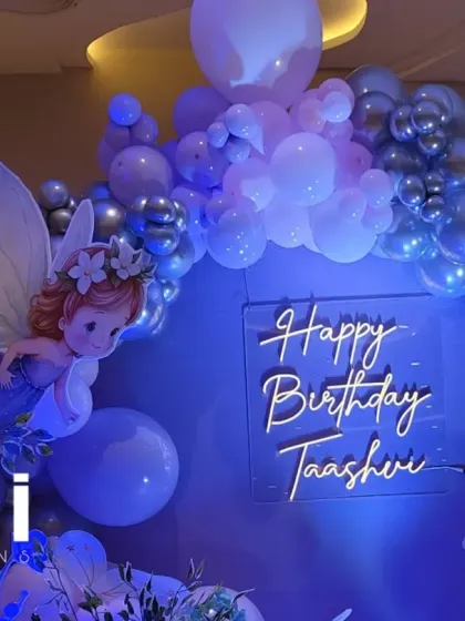 A fairy-themed birthday setup with a character cutout, neon sign, and a silver and purple balloon arch.
