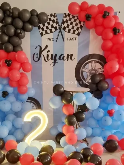 A 'Two Fast' racing-themed second birthday decoration. The setup includes a car-themed backdrop, a balloon arrangement in red, blue, and black, and a light-up neon number '2'.