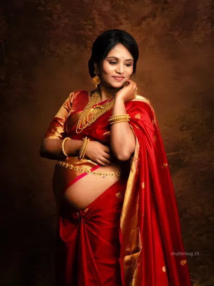 A graceful solo portrait of the mom-to-be, showcasing the beautiful draping of the red silk saree over her baby bump. The intricate gold jewelry completes this elegant, traditional look.