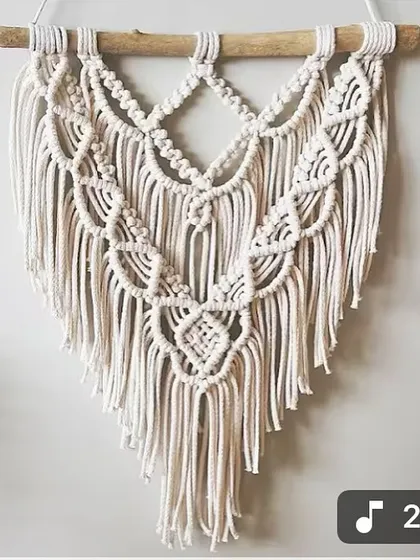 A beautiful, handcrafted macrame piece with layered patterns and a gentle V-shape. It's made from natural cotton cord and hung on a simple wooden dowel.