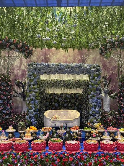A whimsical and enchanting stage design with a backdrop of blue and purple flowers, tree-like props, and a ceiling of hanging greenery.
