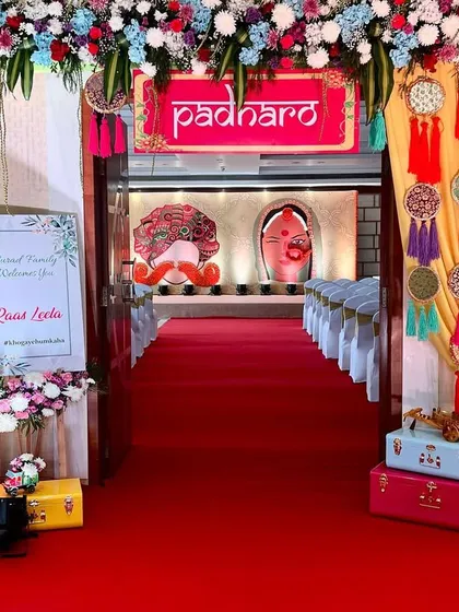 A vibrant and festive entrance for a 'Raas Leela' themed event. A bright red carpet leads guests through a floral archway towards a stage with traditional motifs.