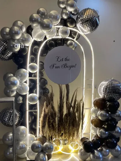 A very glamorous and modern party setup. This features a lit archway filled with pampas grass, surrounded by black, silver, and chrome balloons and disco ball accents for a nightclub vibe.