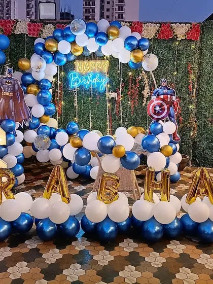 A superhero-themed birthday party featuring both Batman and Captain America. The setup includes a balloon ring against a green wall, character cutouts, a 'Happy Birthday' neon sign, light-up number '6', and balloon letters spelling the name 'PRAHAV'.