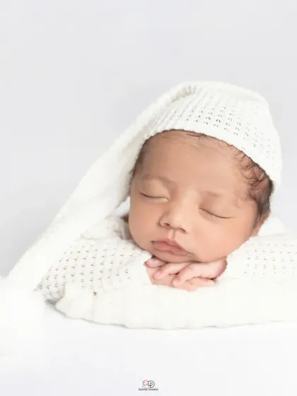 A pure and simple newborn portrait against a clean white background. The baby is dressed in a white knitted outfit and sleepy hat, showcasing their angelic features.