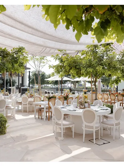 An elegant outdoor dining setup with classic white tables and chairs, surrounded by lush greenery. The setting is perfect for a sophisticated and relaxed wedding luncheon.