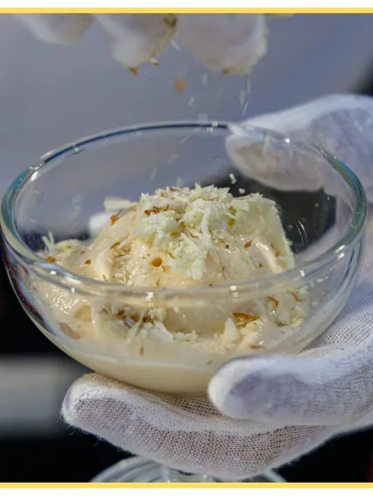 The finishing touch on a custom ice cream creation. A gloved hand sprinkles white chocolate shavings over a scoop of ice cream.